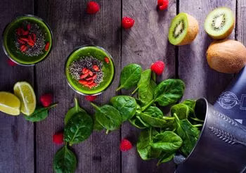 na drewnianym stole dwa zielone koktajle ozdobione superfoods, warzywami i owocami: jagodami goji, liśćmi szpinaku, malinami, kiwi, limonką 
