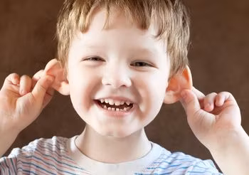 Uśmiechnięty, kilkuletni chłopiec trzymający sie obiema rękami za uszy. 
