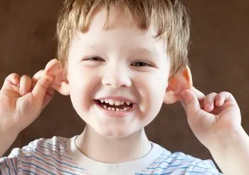 Uśmiechnięty, kilkuletni chłopiec trzymający sie obiema rękami za uszy. 