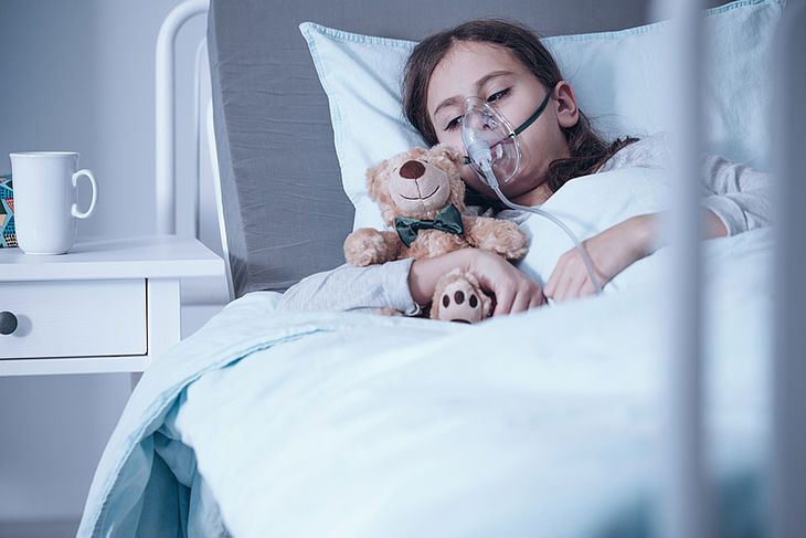 Dziewczynka z aparatem tlenowym leży na łóżku i przytula misia