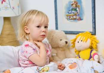 Mała dziewczynka siedząca na łóżku w otoczeniu lalek i trzymająca się za gardło 