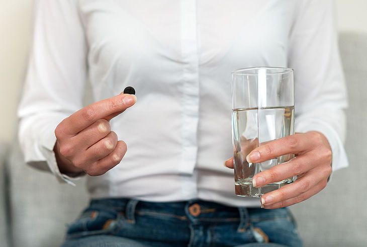 Kobbieta trzyma w jednej dłoni tabletkę węgla aktywnego, w grugiej szklankę wody.