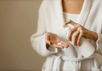 Kobieta w białym szlafroku trzyma w dłoni butelkę z balsamem-emolientem.
