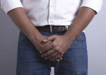 Mężczyzna w dżinsach i koszuli trzymający dlonie w okolicy krocza. 