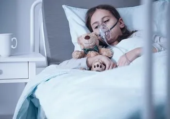 Dziewczynka z aparatem tlenowym leży na łóżku i przytula misia