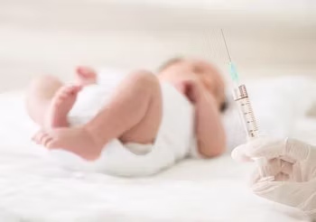 Na pierwszym planie ręka lekarza z przygotowaną szczepionką, na drugim planie - niemowlę