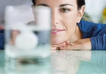 Na pierwszym planie szklanka z wodą, w której rozpuszcza się aspiryna, na drugim planie młoda kobieta. 