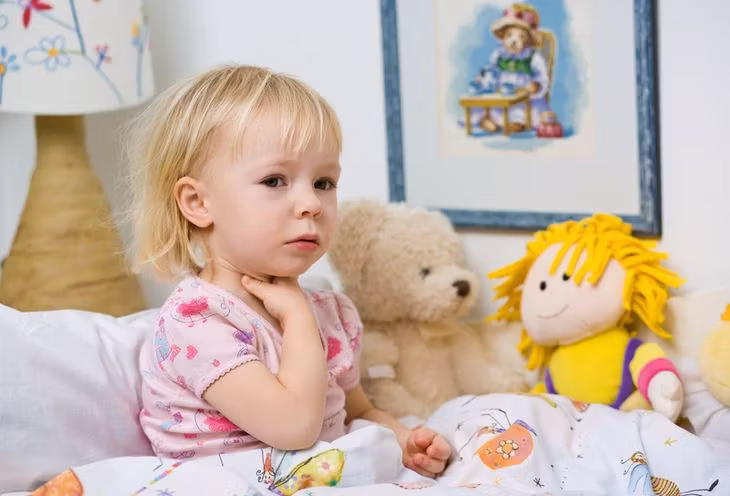 Mała dziewczynka siedząca na łóżku w otoczeniu lalek i trzymająca się za gardło 